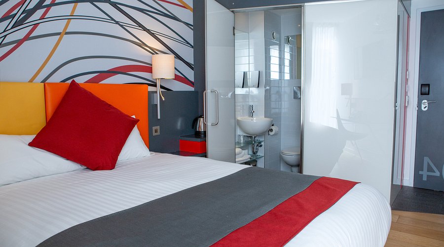 Small uniform hotel room with a fitted bed, desk shelf and window blind